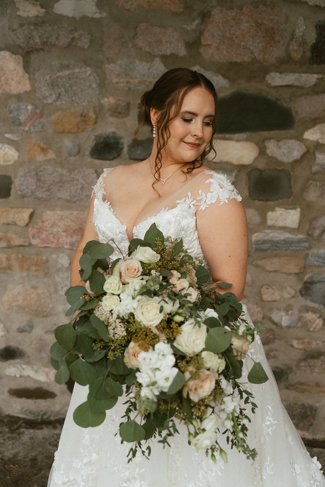 Bride holding a cascading bouquet of white roses and eucalyptus against a stone wall