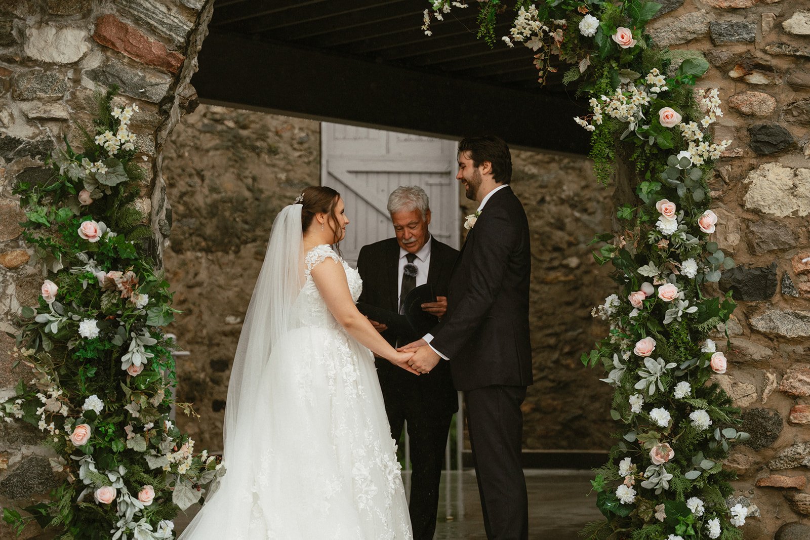 Bride and groom exchanging vows beneath a floral arch against a stone wall